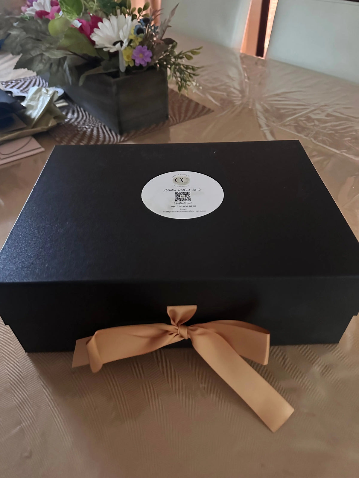 A black rectangular gift box with a round white label on the side and a beige satin ribbon tied in a bow, photographed on a beige surface with flowers partially visible at the lower left.