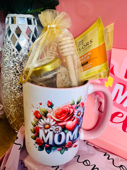 Close-up of a white ceramic mug featuring a colorful floral design and the word "MOM." Inside the mug is a gold mesh gift bag containing a small honey jar and a wooden honey dipper, tucked next to two yellow Taylors of Harrogate Mandarin & Ginger tea sachets.