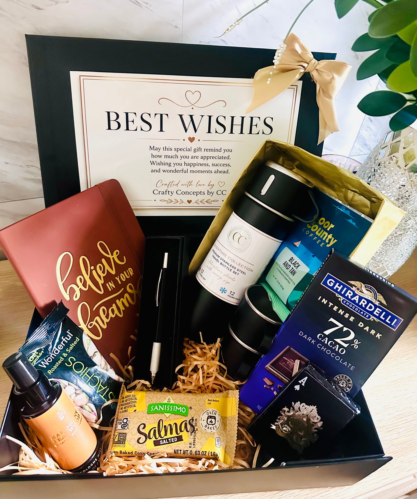 A curated gift basket arrangement in a black tray with shredded paper filler, featuring a brown notebook with gold script, a black pen, a white bottle, a blue and green packaged item, a black candle, a yellow packet of Salty Snacks, a dark chocolate bar, and a large black gift box behind it with a gold bow and a 'BEST WISHES' card.