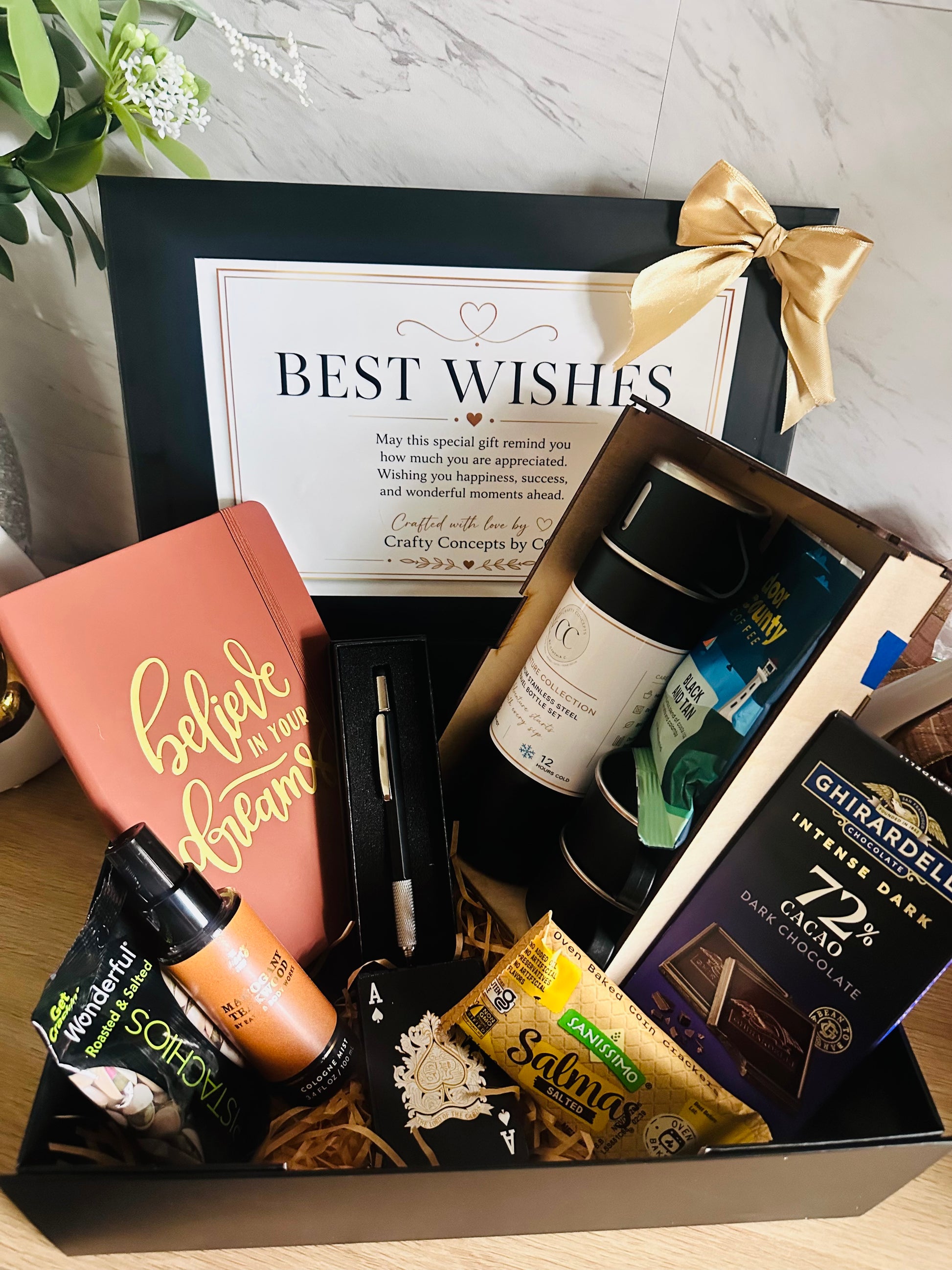 A curated gift box filled with assorted items, including a pink notebook, a bottle, a pen, a tube of soap, a bag of Salty's Salmon, a bag of Ghedarelbe 72% dark chocolate, and a black gift box with a gold bow and a 'Best Wishes' card.