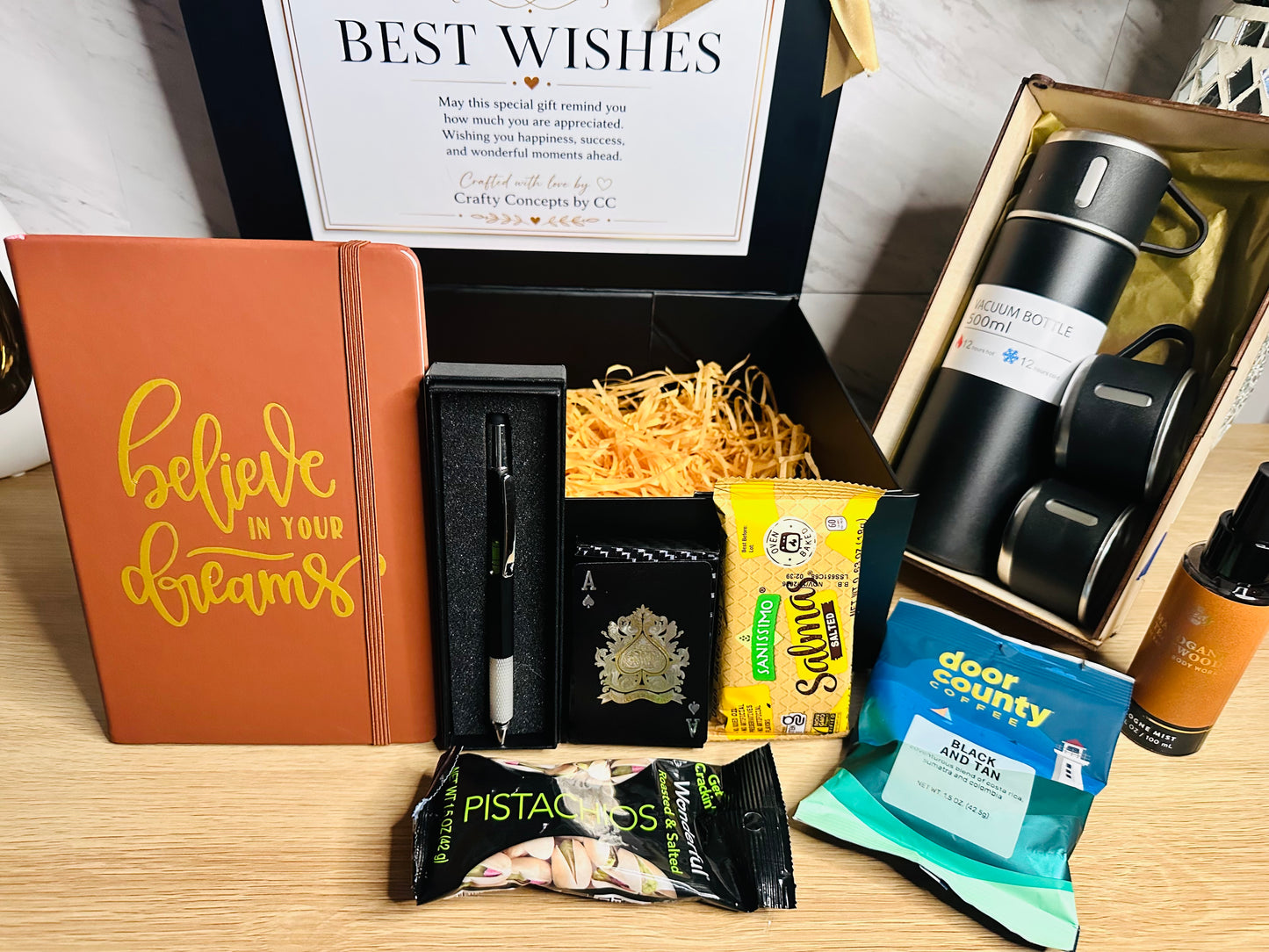 A curated gift box set on a wooden table, featuring a terracotta notebook with gold script reading 'believe in your dreams', a black pen in a black case, a black wallet with a gold crest, a yellow bag of Slim's chips, a blue bag of Door County Blue Cheese, a chocolate-covered pistachio snack, a black travel bottle, and a black insulated tumbler in its box.
