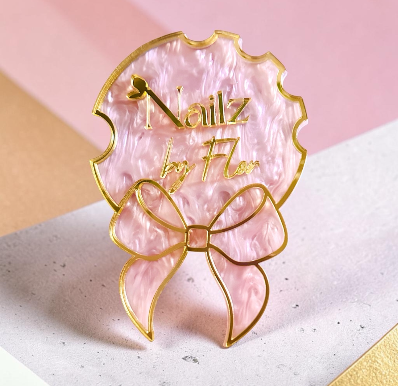 A pink marbled nail art fan-shaped pin with a gold bow and the text 'Nailz by Nae', shown on a pink and beige background.