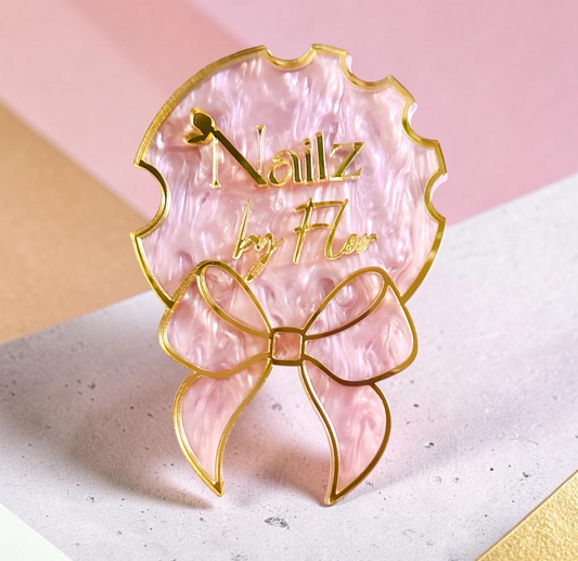 A pink marbled nail art fan-shaped pin with a gold bow and the text 'Nailz by Nae', shown on a pink and beige background.