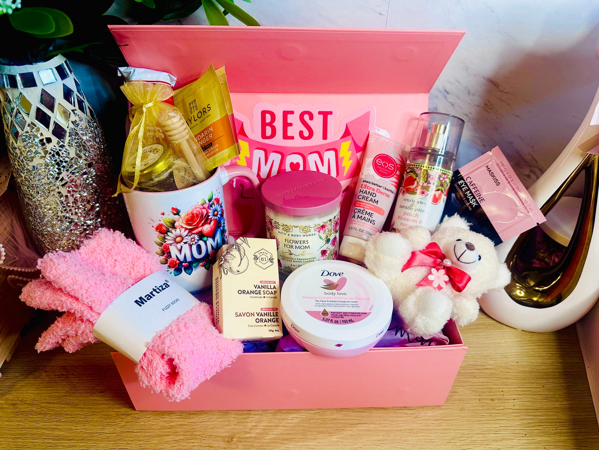 A pink "Best Mom Ever" gift box filled with self-care items including a floral "MOM" mug, a small white teddy bear, Dove body cream, pink fuzzy socks, a vanilla orange soap bar, EOS hand cream, and a Bath & Body Works fragrance mist. The box is set against a marble background next to a decorative mosaic vase.