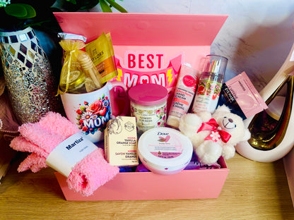 A pink "Best Mom Ever" gift box filled with self-care items including a floral "MOM" mug, a small white teddy bear, Dove body cream, pink fuzzy socks, a vanilla orange soap bar, EOS hand cream, and a Bath & Body Works fragrance mist. The box is set against a marble background next to a decorative mosaic vase.