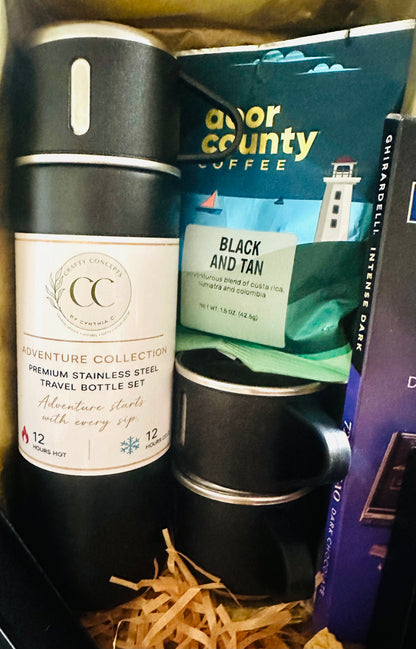 A travel set featuring a black stainless steel bottle with a white label, a black travel mug, and a bag of Door County Coffee Black and Tan, arranged in a gift box with shredded paper.