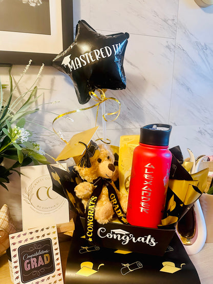 Graduation gift basket with red laser engraved water bottle, plush teddy bear in grad cap, white chocolates and black Mastered It star balloon in a Congrats gift box