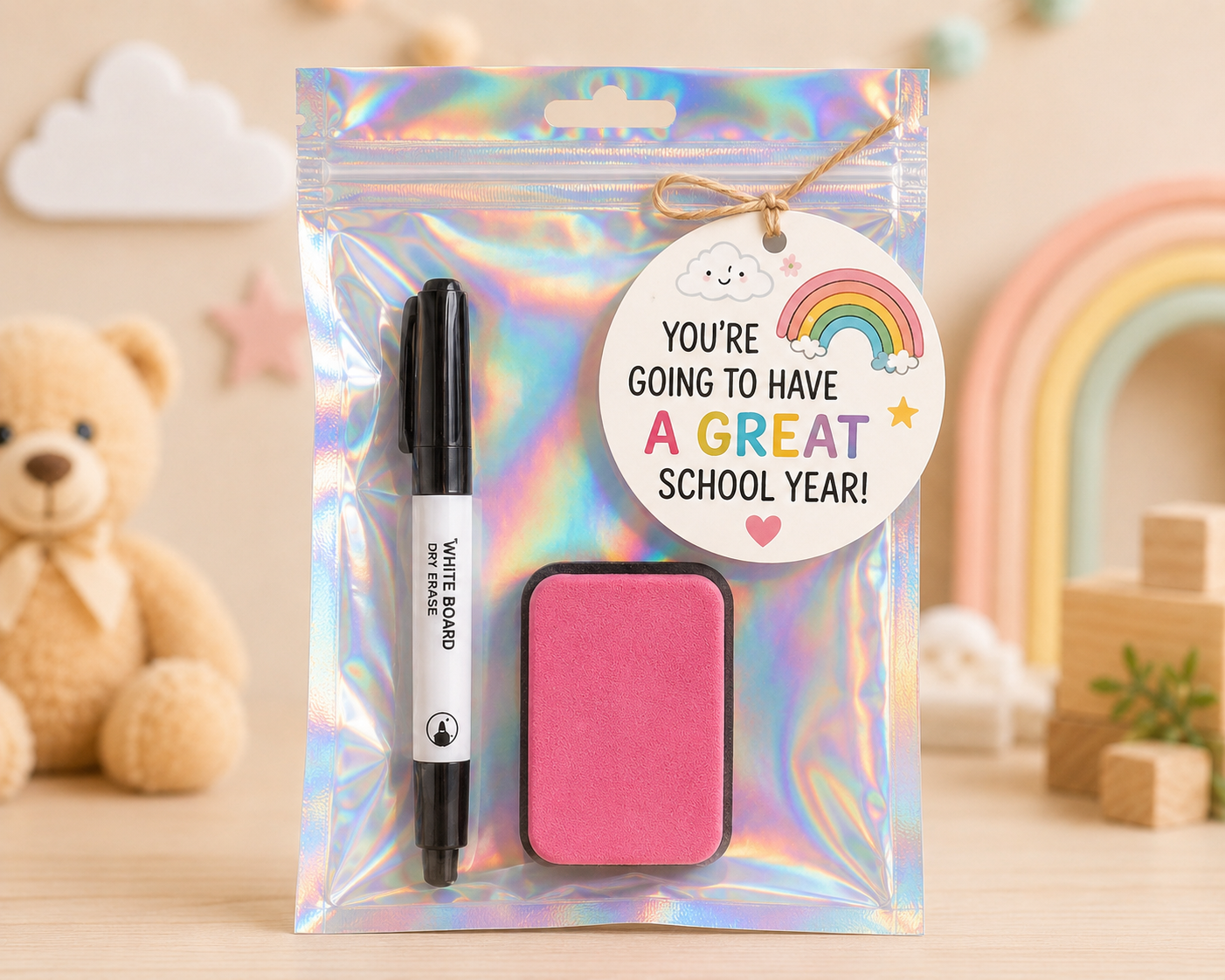 A holographic zip pouch gift set containing a black whiteboard pen, a pink sticky note pad, and a round white hanging tag that reads 'YOU'RE GOING TO HAVE A GREAT SCHOOL YEAR!' with a rainbow and cloud illustration. The pouch is photographed on a light tabletop with a blurred teddy bear, rainbow, and blocks in the background.