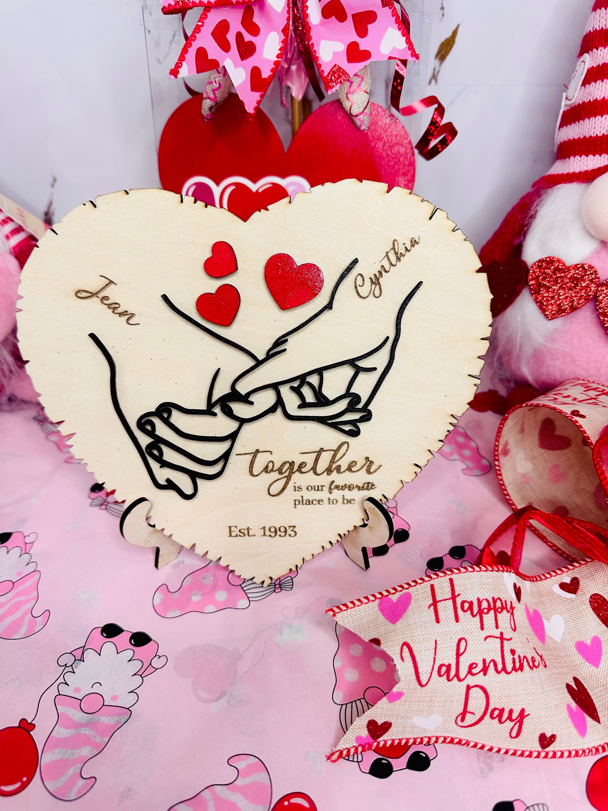 Personalized heart-shaped wooden plaque featuring a black and white "pinky promise" illustration, custom engraved with the names Joan and Cynthia and "Est. 1993." The board includes the quote "Together is our favorite place to be" and is surrounded by red Valentine's Day decor.