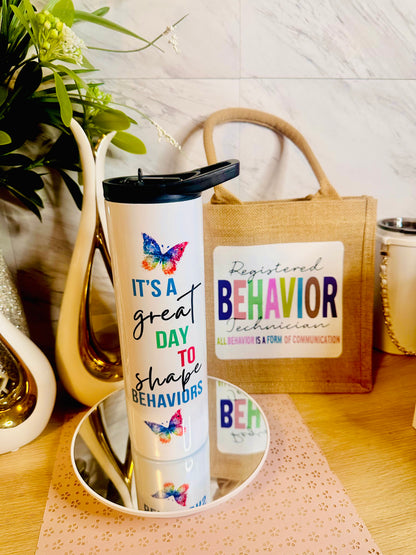 A white insulated tumbler with a black lid stands on a mirrored tray on a wooden table. The tumbler has colorful butterfly graphics and the text 'IT'S A great DAY TO shape BEHAVIORS'.