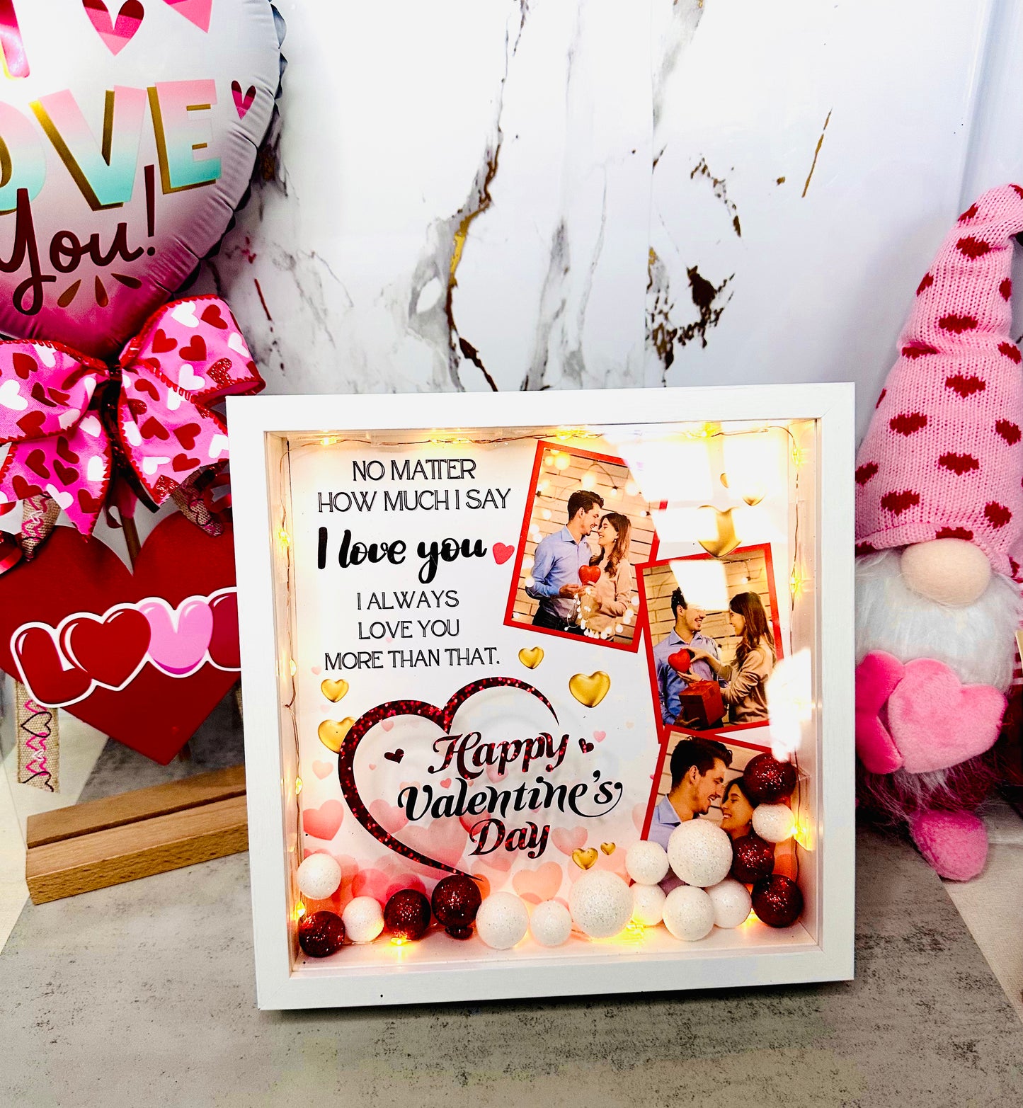 A white personalized shadow box for Valentine's Day featuring two custom couple photos, warm LED fairy lights, and red and white glitter sphere fillers with a romantic quote printed on the glass.