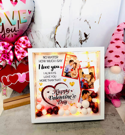 A white personalized shadow box for Valentine's Day featuring two custom couple photos, warm LED fairy lights, and red and white glitter sphere fillers with a romantic quote printed on the glass.