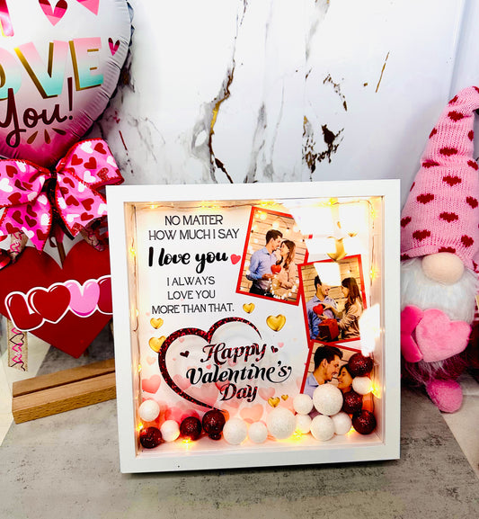 A white personalized shadow box for Valentine's Day featuring two custom couple photos, warm LED fairy lights, and red and white glitter sphere fillers with a romantic quote printed on the glass.