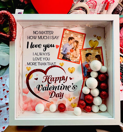 A white personalized shadow box for Valentine's Day featuring two custom couple photos, warm LED fairy lights, and red and white glitter sphere fillers with a romantic quote printed on the glass.