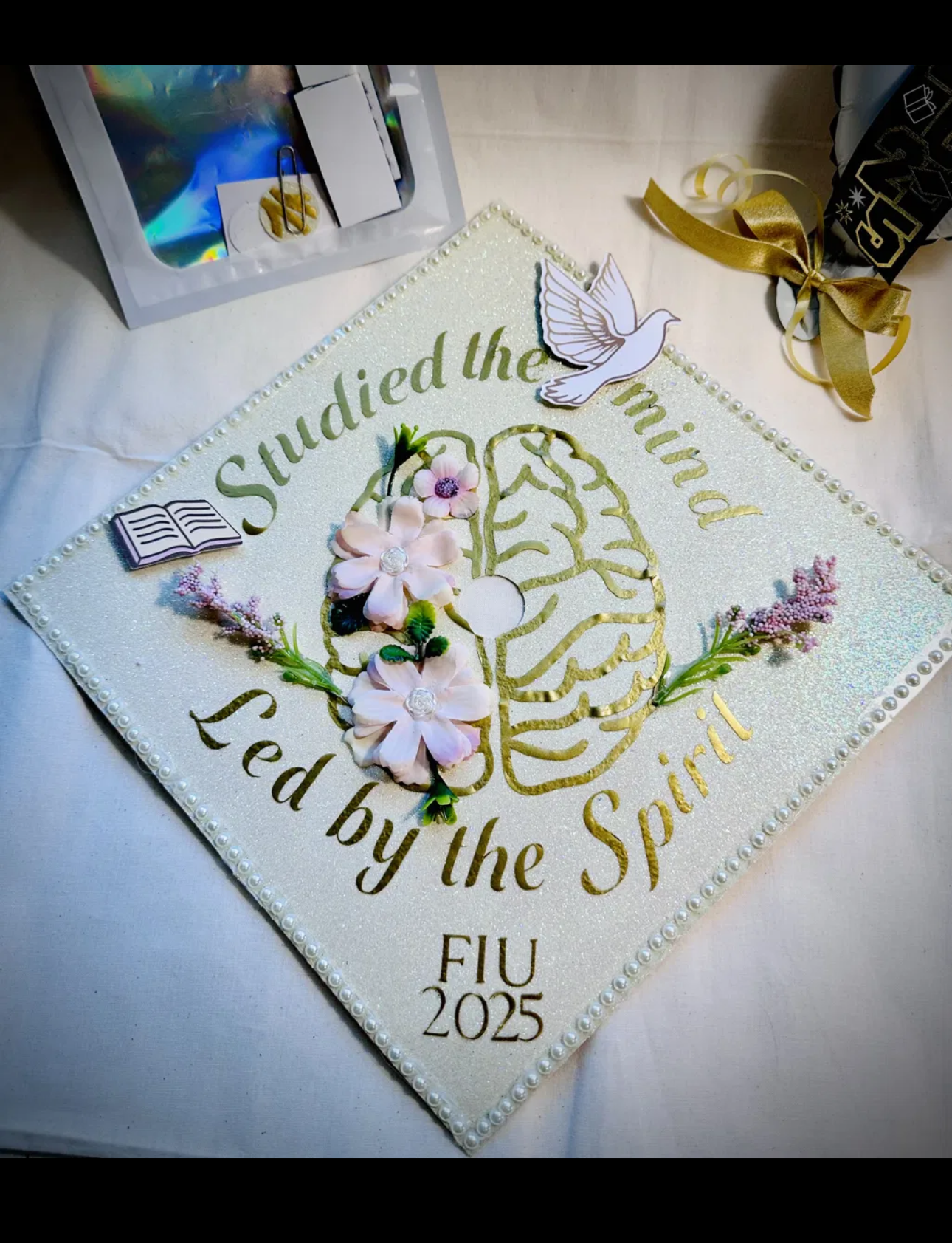 Custom graduation cap topper with white glitter background, gold brain design, pink flowers, pearls and dove reading Studied the Mind Led by the Spirit FIU 2025