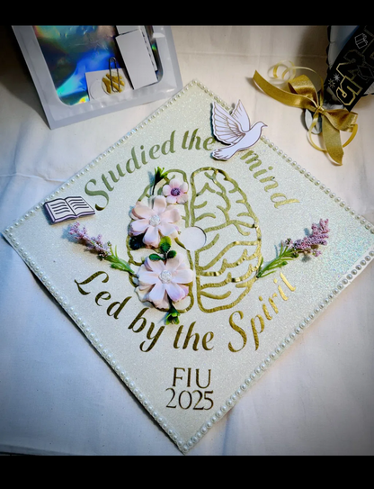 Custom graduation cap topper with white glitter background, gold brain design, pink flowers, pearls and dove reading Studied the Mind Led by the Spirit FIU 2025